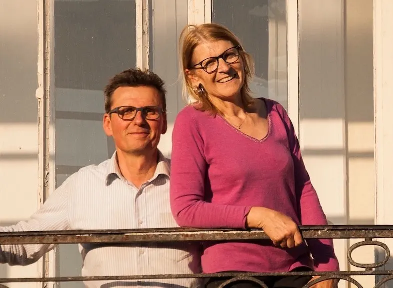 Les propriétaires de La Maison M, souriants, accoudés à une balustrade en fer forgé au soleil
